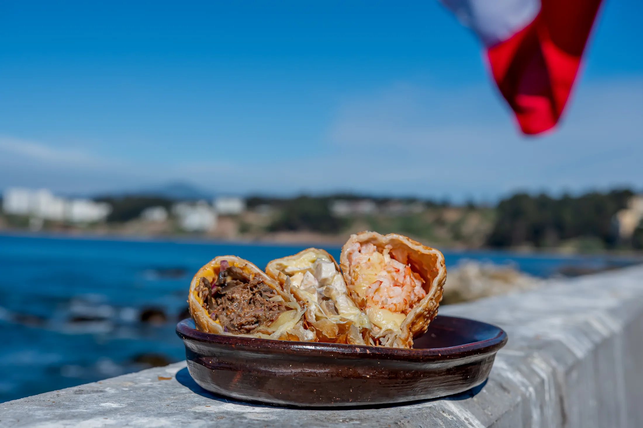Empanadas abiertas sobre una fuente de greda frente al mar de Horcón, con la bandera chilena flameando.